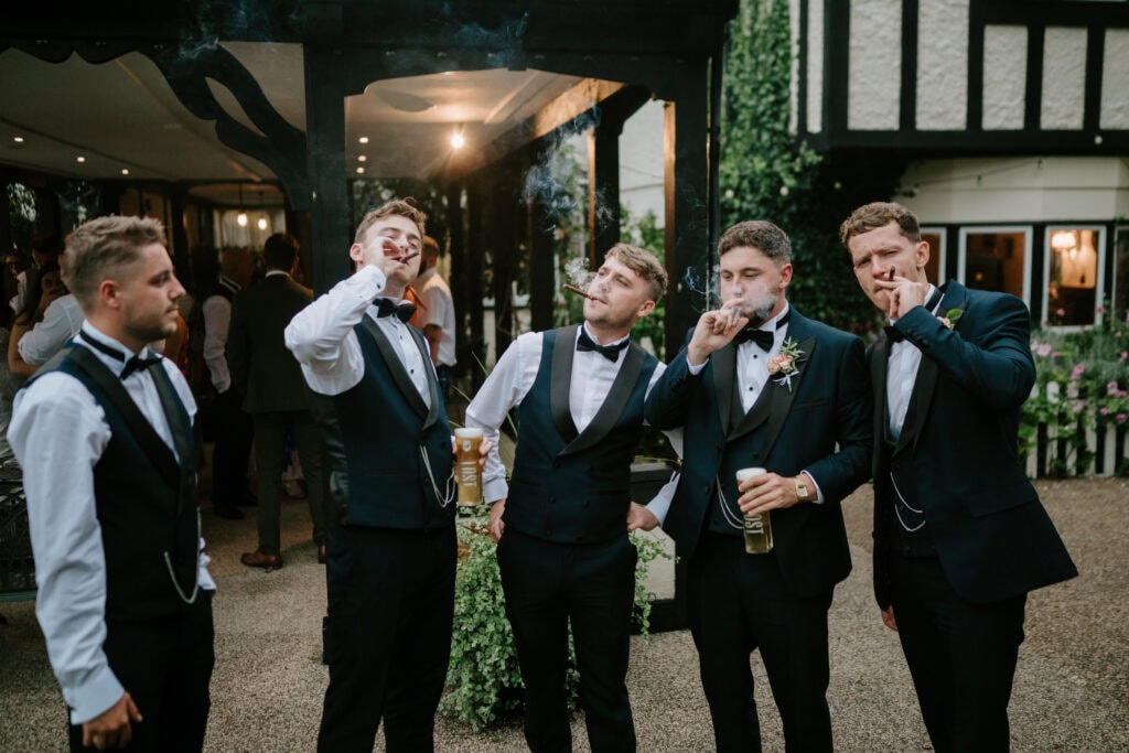 A group of five men in formal attire stand outside at a wedding in Kent, smoking cigars and holding drinks. They are dressed in black suits with black bow ties. The background shows Hayne House with its Tudor-style facade and greenery around the entrance. Image by Pearce Wedding Photography.