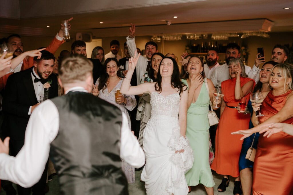 A joyful bride in a white dress and groom in a tuxedo vest dance together at their wedding reception at Hayne House in Kent. They are surrounded by cheering and smiling guests, who are also dancing and holding drinks, creating a lively and celebratory atmosphere. Image by Pearce Wedding Photography.