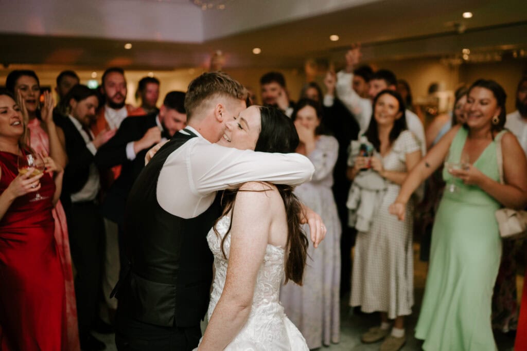 A bride and groom share a kiss on the dance floor at Hayne House in Kent, surrounded by cheering guests in a lively wedding reception. The bride, in her white gown, and the groom, in his black tuxedo, bask in the joyous celebration. Image by Pearce Wedding Photography.