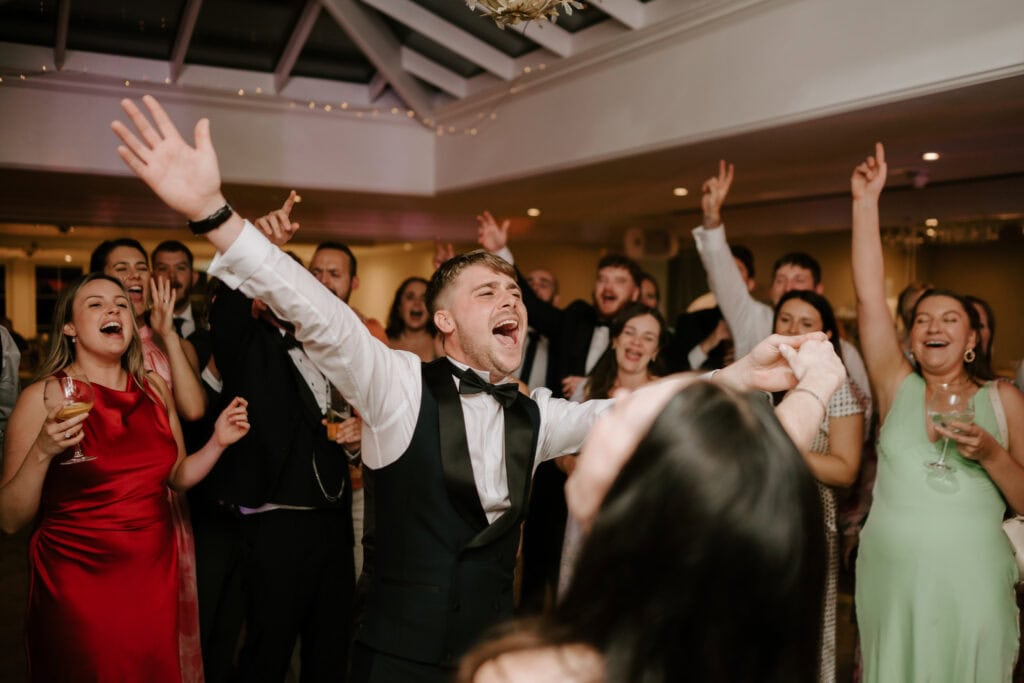 A joyful group of people are dancing and singing at a lively indoor wedding celebration at Hayne House in Kent. A man in a suit is in the foreground with his arms raised in excitement, surrounded by smiling and cheering individuals dressed in formal attire. Everyone appears to be having a great time. Image by Pearce Wedding Photography.