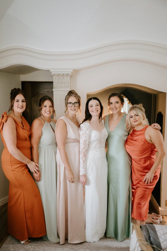 A bride stands in the center wearing a white wedding dress, surrounded by five bridesmaids in various pastel and bright dresses. They are all smiling and posing inside a beautifully decorated room at Hayne House, with an elegant archway in the background. Image by Pearce Wedding Photography.