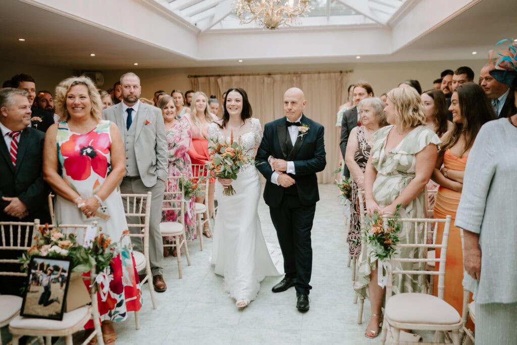 A bride in a white dress walks down the aisle at Hayne House, holding a bouquet and accompanied by the father of the bride in a suit. Guests on both sides smile and watch. The room is decorated with flowers, and natural light streams in through a skylight. Image by Pearce Wedding Photography.