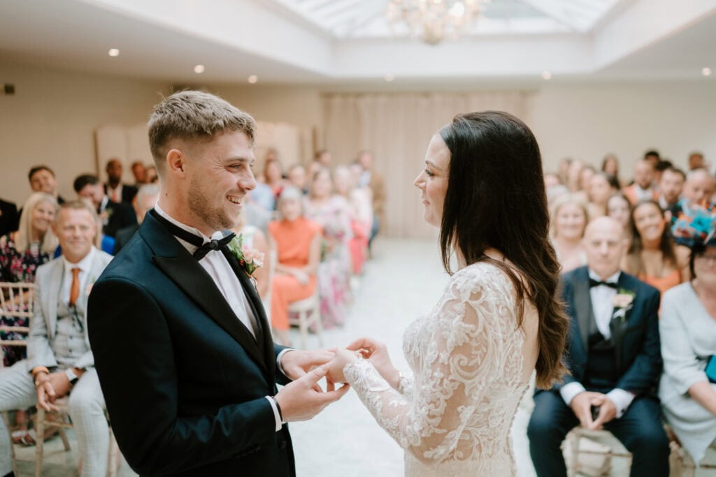 A bride and groom stand facing each other, exchanging rings during their wedding ceremony at Hayne House in Kent. The bride is wearing a lace dress while the groom is in a black tuxedo. Guests are seated behind, attentively watching the couple in a bright, airy room with a glass ceiling. Image by Pearce Wedding Photography.