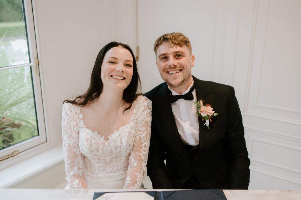 A happy couple sits together, smiling brightly. The woman wears a white lace wedding dress, and the man is in a black suit with a bow tie and a pink boutonniere. They are indoors at Hayne House in Kent, with a white wall and window in the background, capturing the perfect wedding moment. Image by Pearce Wedding Photography.