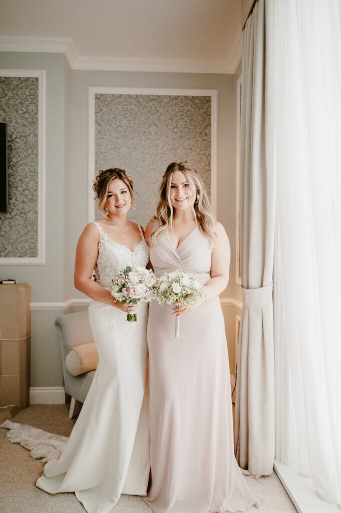 Two women smile while posing indoors beside a curtain at the Hythe Imperial Hotel. One wears a white wedding gown with lace details and holds a small bouquet of flowers, while the other is in a blush pink bridesmaid dress, also holding a bouquet. The background features light green walls and upholstered furniture, perfectly captured by the Hythe wedding photographer. Image by Pearce Wedding Photography.