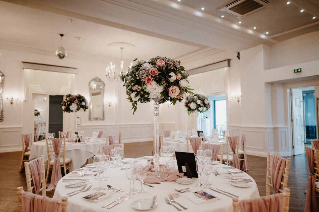 A beautifully decorated banquet hall at the Hythe Imperial Hotel features round tables set with elegant tableware, glasses, and napkins. Each table showcases a tall floral centerpiece with pink and white flowers. Soft lighting and chandeliers create a warm ambiance, perfect for a Hythe wedding photographer to capture. Image by Pearce Wedding Photography.