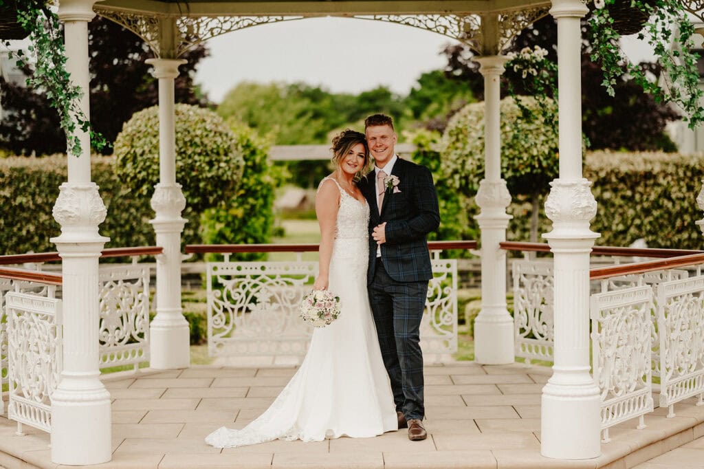 A bride and groom stand happily under a decorated gazebo adorned with green vines. The bride wears a white wedding dress and holds a bouquet of flowers, while the groom is dressed in a dark checkered suit. They are outdoors, surrounded by greenery at the picturesque Hythe Imperial Hotel. Image by Pearce Wedding Photography.