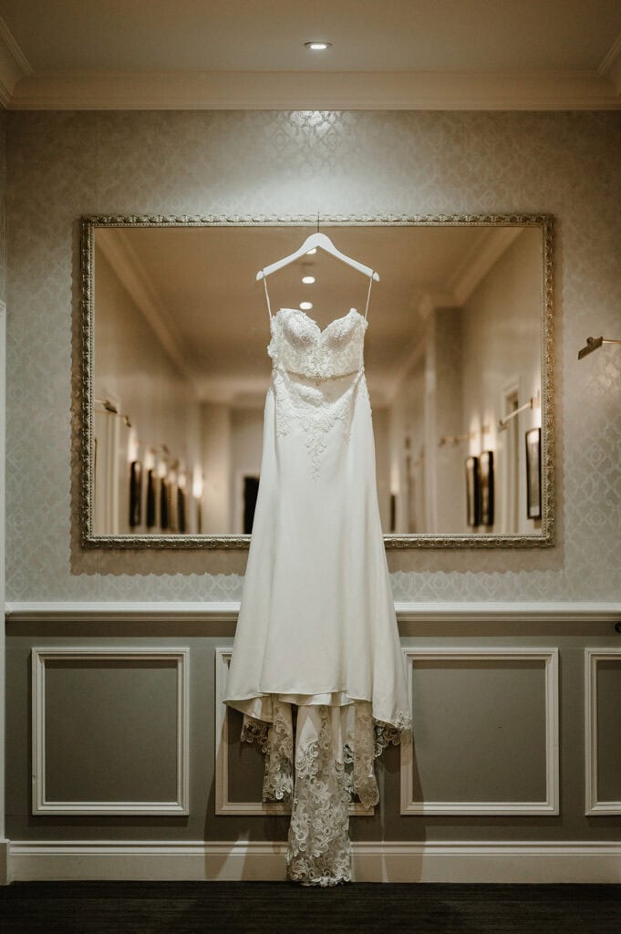 A strapless, white wedding dress with lace details hangs on a hanger in front of a large framed mirror. The dress features a fitted bodice and a flowing train with intricate lace at the bottom. The setting, reminiscent of the Hythe Imperial Hotel, is a hallway with elegant decor and soft lighting. Image by Pearce Wedding Photography.