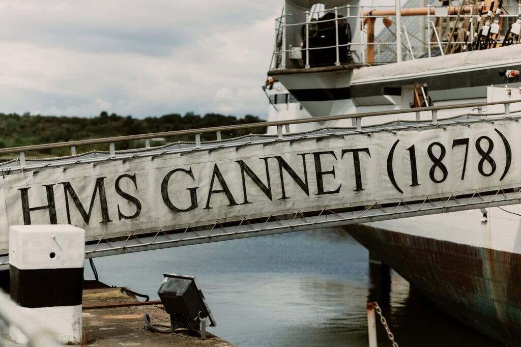 A close-up view of a gangway with the inscription "HMS GANNET (1878)" leading to a historic naval ship docked at Chatham Dockyard. The background includes a calm water body and partial ship detailing with rigging, evoking both maritime and historical atmospheres suitable for a Medway wedding photographer. Image by Pearce Wedding Photography.