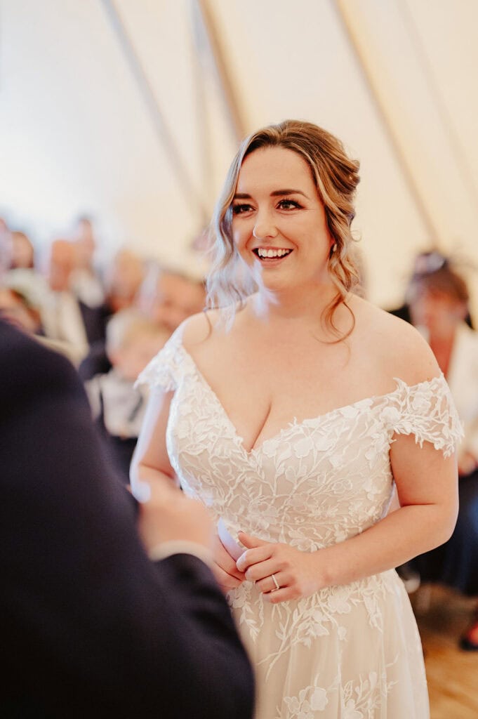 A bride in a white lace wedding dress smiles during a wedding ceremony inside a tent. She has long hair styled in loose waves and is holding a ring, captured beautifully by the Medway wedding photographer. People are seated in the background, slightly out of focus. Image by Pearce Wedding Photography.