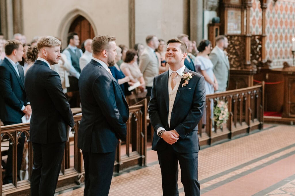 A groom stands at the altar of Rochester Cathedral, smiling and looking upward while wearing a dark suit with a light vest and tie. Groomsmen stand nearby in matching suits. In the background, guests seated in pews observe the ceremony surrounded by the cathedral's ornate decorations, captured beautifully by a Medway wedding photographer. Image by Pearce Wedding Photography.