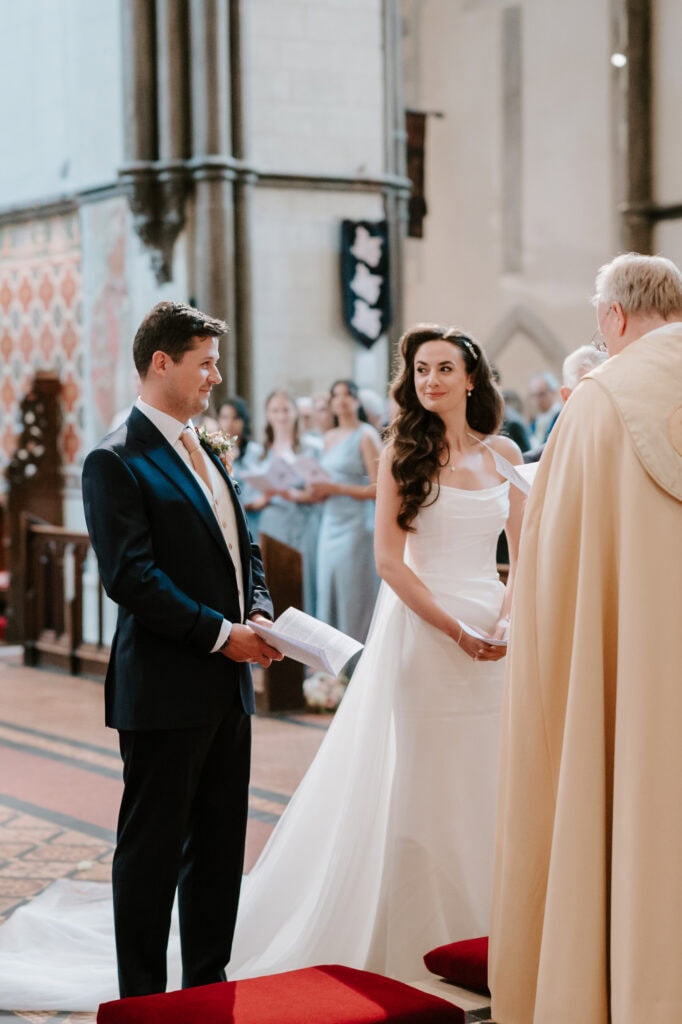 A couple stands at the altar during a church wedding ceremony. The groom, in a dark suit and light vest, smiles and holds a booklet. The bride, in a white dress with a long train, gazes at the groom. A clergyman in a cream robe stands before them as the Medway wedding photographer captures every moment. Image by Pearce Wedding Photography.