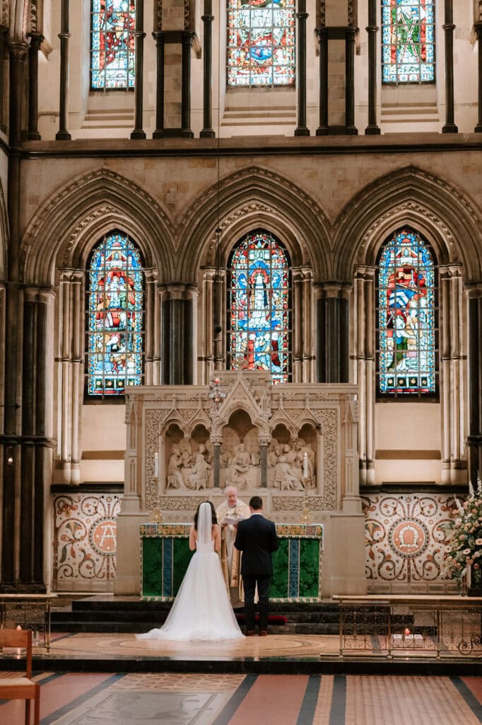 A bride and groom stand at the altar in Rochester Cathedral, with ornate stained glass windows behind them. The bride is wearing a white gown and veil, while the groom is in a dark suit. The priest, in light robes, conducts the ceremony in front of a detailed stone backdrop captured by their Medway wedding photographer. Image by Pearce Wedding Photography.