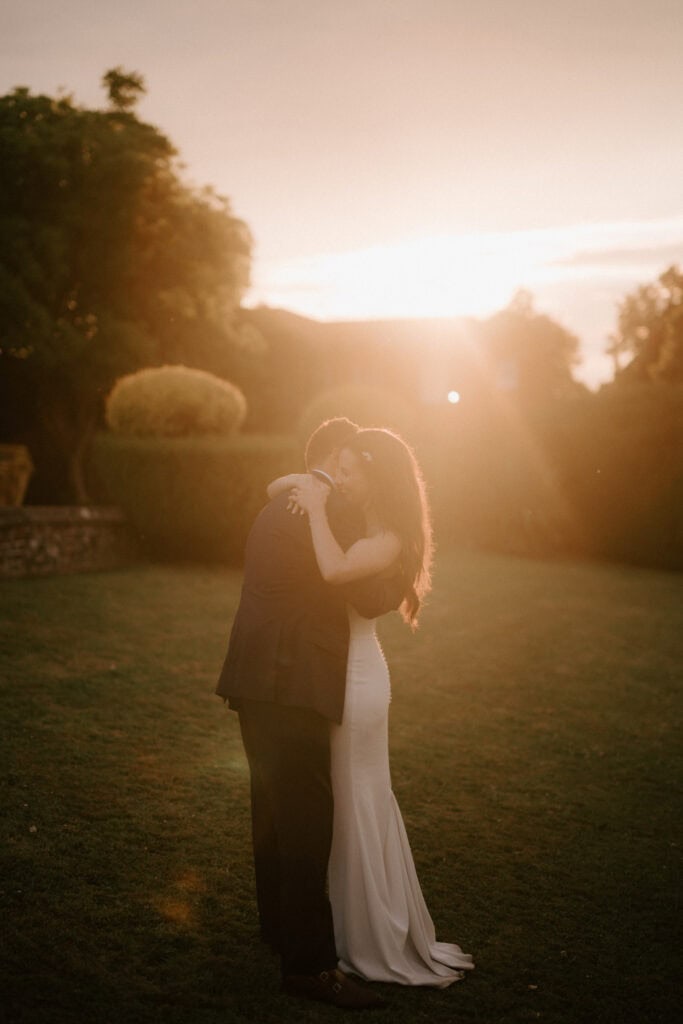 A couple embraces on a grassy field during sunset, surrounded by lush greenery. Captured by a talented Kent wedding photographer, the woman in a long, elegant dress and the man in a suit are bathed in the sun’s warm glow, creating an enchantingly romantic atmosphere. Image by Pearce Wedding Photography.