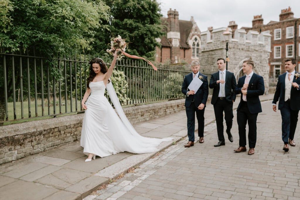 Bride walks joyfully with groomsmen on cobbled street.