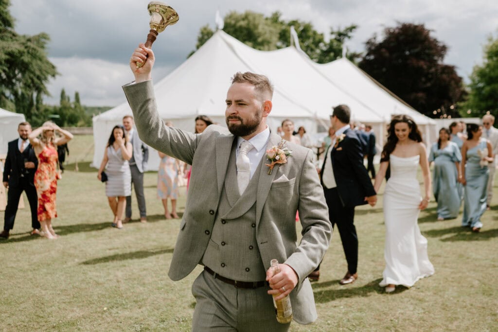 Man in suit with bell, outdoor wedding celebration.