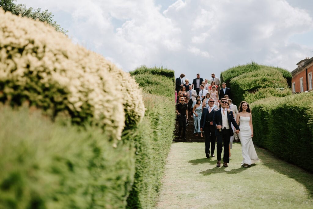 At a picturesque Kent wedding venue, a bride and groom stroll down a path through manicured hedges, followed by a group of smiling guests on a sunny day. Image by Pearce Wedding Photography.