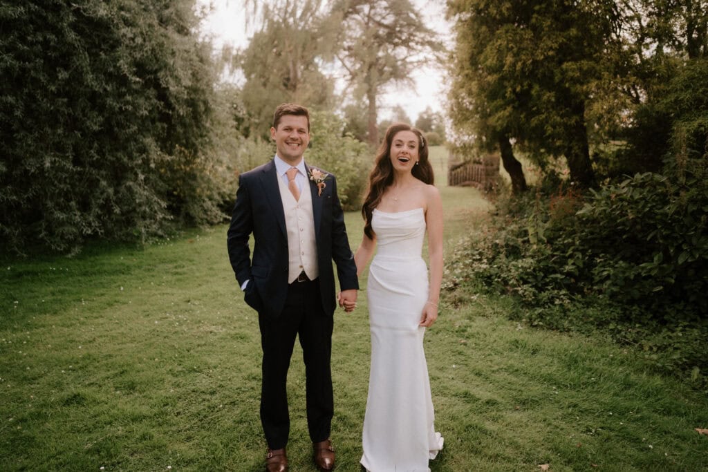 A bride and groom, captured in exquisite wedding photography, stand together on a grassy path surrounded by trees. The groom wears a navy suit, and the bride is radiant in her white dress. Smiling and holding hands, they are framed by a lush, green backdrop. Image by Pearce Wedding Photography.