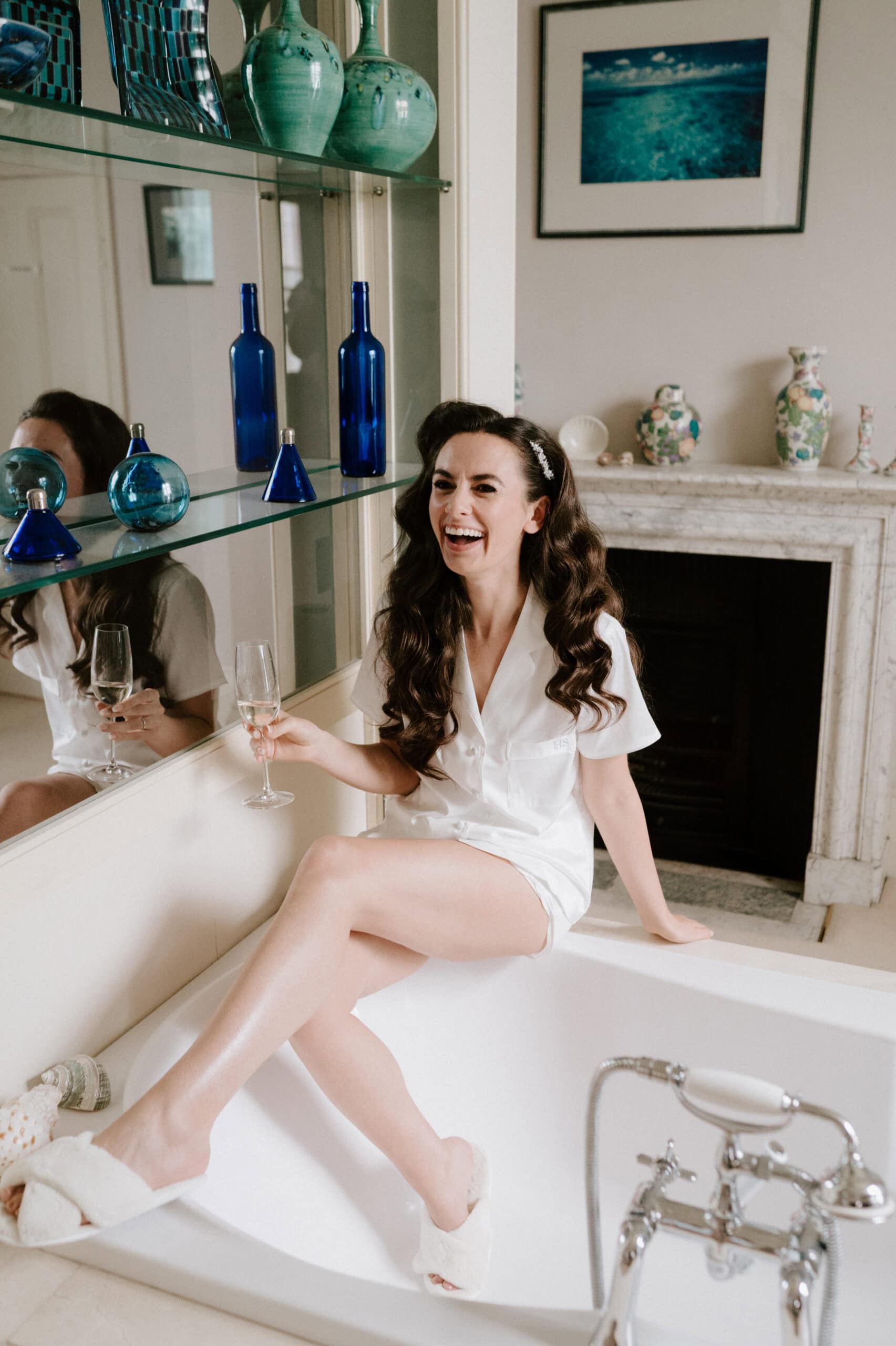 A fun bride in Bath, dressed in white pyjamas and slippers, sits on the tub’s edge with a glass of wine, laughing. The bathroom is adorned with blue and green vases on glass shelves and a cosy fireplace in the background. Image by Pearce Wedding Photography.