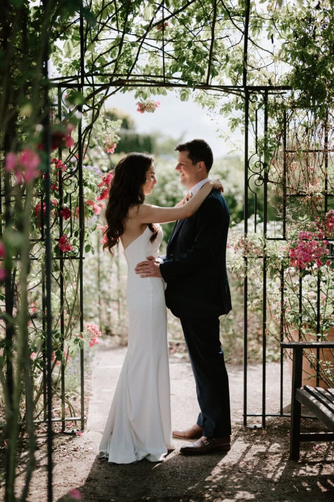 Couple in garden archway, embracing on wedding day.