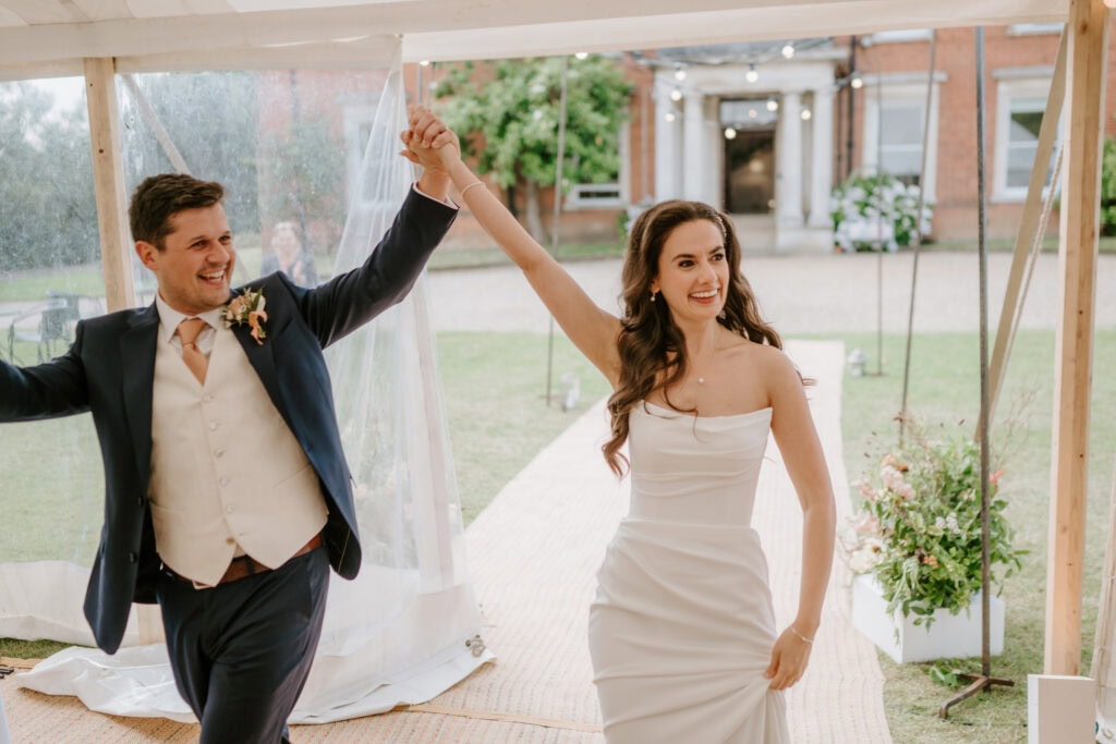 A smiling couple, dressed in wedding attire, dances joyfully under a tent at their Mount Ephraim wedding. The groom is in a navy suit and vest, while the bride dazzles in her white strapless gown. Flowers and a charming red-brick building set the perfect Kent backdrop. Image by Pearce Wedding Photography.