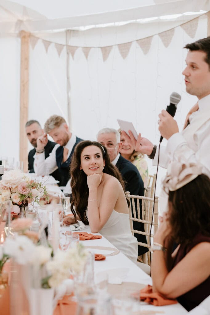 At a charming Kent wedding, the bride sits at a reception table, gazing up at the speech-giver with microphone in hand. Guests in formal attire smile warmly around her. The table is gracefully adorned with flowers, and a white draped tent creates a soft backdrop reminiscent of Mount Ephraim's elegance. Image by Pearce Wedding Photography.