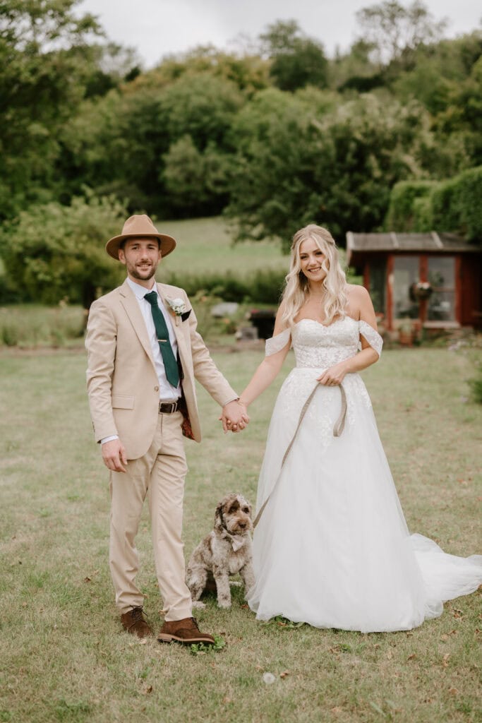 A couple in formal attire stands hand in hand on a grassy field, smiling for their Kent wedding photographer. The man wears a beige suit and hat, while the woman is radiant in her white wedding dress. Beside them, a small, fluffy dog sits obediently on a leash amid the lush greenery. Image by Pearce Wedding Photography.