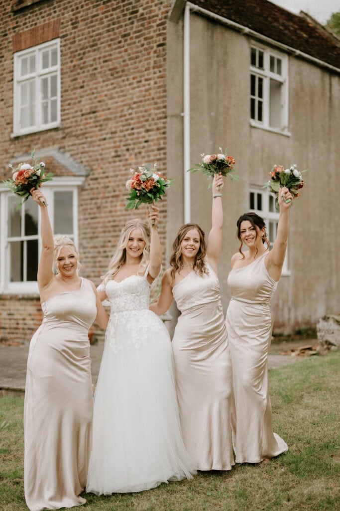 A bride in a white gown and three bridesmaids in matching beige dresses stand outside a brick and stucco house. They are smiling and holding bouquets of flowers, raising them joyfully in the air. Captured by a talented photographer, the image conveys a celebratory and cheerful atmosphere. Image by Pearce Wedding Photography.