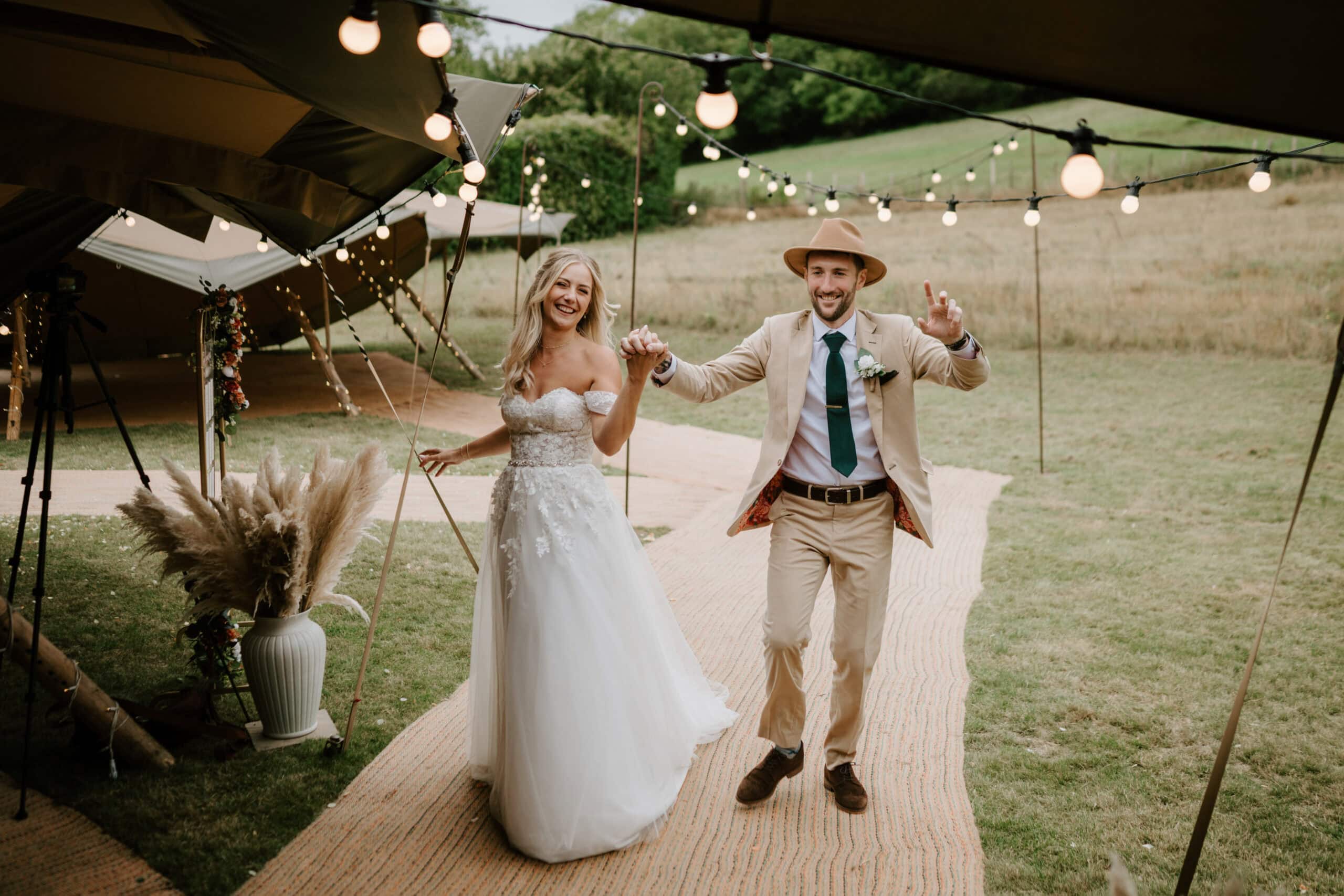 A bride and groom joyfully walk hand in hand under festive string lights, beautifully captured by a kent wedding photographer. The bride wears a white gown, and the groom dons a beige suit with a hat. They are outdoors, surrounded by greenery and a decorated tent. Image by Pearce Wedding Photography.