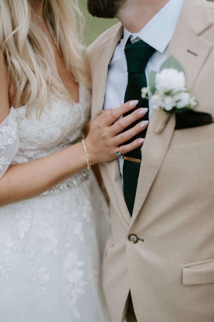 A bride in a lace wedding dress gently places her hand on the chest of a groom wearing a beige suit and dark green tie with a floral boutonnière. Captured by a skilled photographer in Rochester, the intimate moment focuses on their torsos and hand placement. Image by Pearce Wedding Photography.
