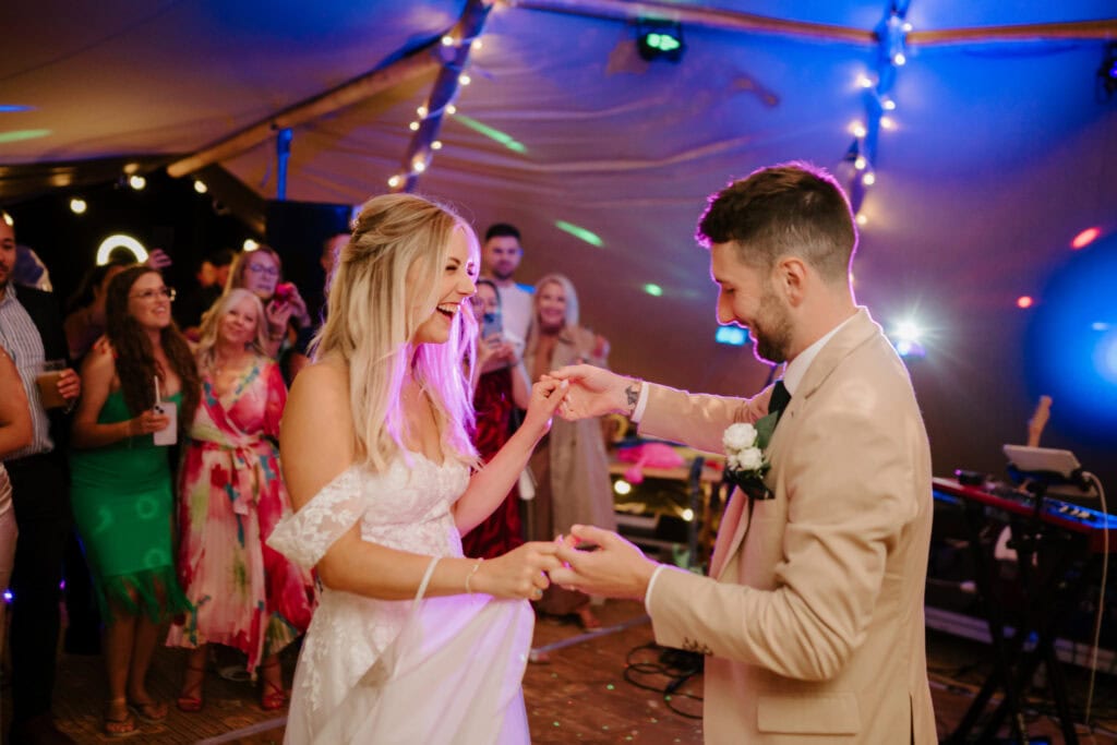 A bride and groom dance joyfully under bright, colorful lights at their wedding reception. The bride wears an off-shoulder white gown, while the groom is in a beige suit. Friends and family in festive attire surround them, celebrating as photographers capture every magical moment of this festival of love. Image by Pearce Wedding Photography.