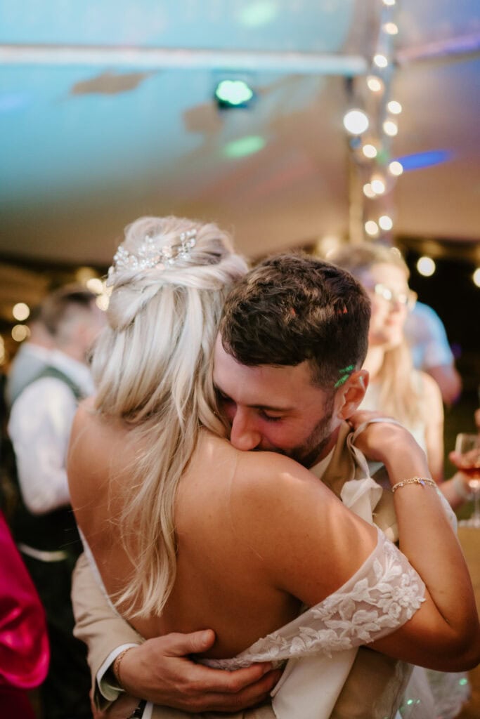 A bride and groom share an intimate embrace during their wedding reception. The bride, wearing a strapless gown, has her back to the camera. The groom holds her tightly, his eyes closed. Twinkling lights create a warm, festive atmosphere reminiscent of Rochester's famed festival in the background. Image by Pearce Wedding Photography.