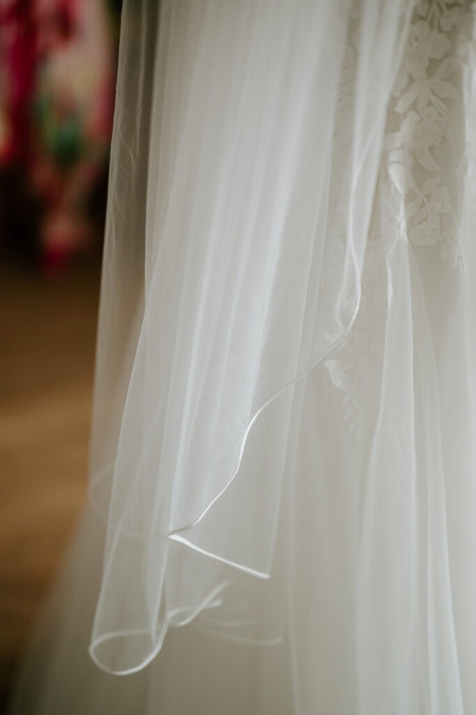 Close-up of a flowing, white bridal veil with delicate lace details. The fabric is sheer and light, softly cascading down with a blurred, colorful background. Captured by a talented Rochester photographer, the image exudes an elegant and romantic atmosphere akin to a dreamy festival scene. Image by Pearce Wedding Photography.