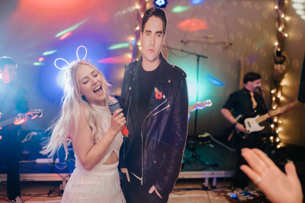 A festival scene unfolds as a woman wearing glowing bunny ears sings into a microphone while hugging a life-size cardboard cutout of a man in a leather jacket. Behind her, colorful lights illuminate the stage where the band plays. A photographer captures this whimsical moment amidst the festivities. Image by Pearce Wedding Photography.