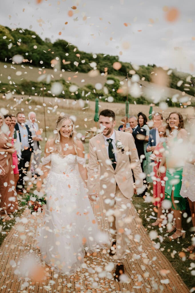 At their Rochester wedding, a bride and groom walk down an outdoor aisle, smiling and holding hands. They're surrounded by guests who are throwing flower petals. The bride wears a white dress, and the groom is in a beige suit. The setting is verdant with trees and hills in the background. Image by Pearce Wedding Photography.