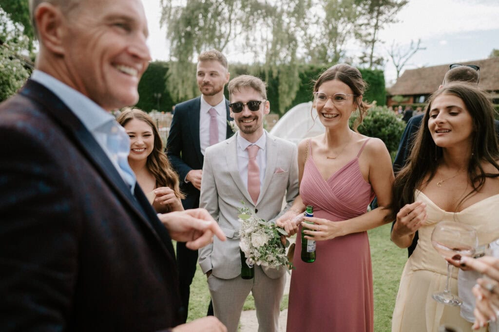 A group of well-dressed people, smiling and engaging in conversation at an outdoor event, sets a charming scene. One person in the foreground, holding a drink, appears to be the focus of the conversation. The casual setting of Winters Barns with greenery and rustic buildings hints at a picturesque Kent wedding ambiance. Image by Pearce Wedding Photography.