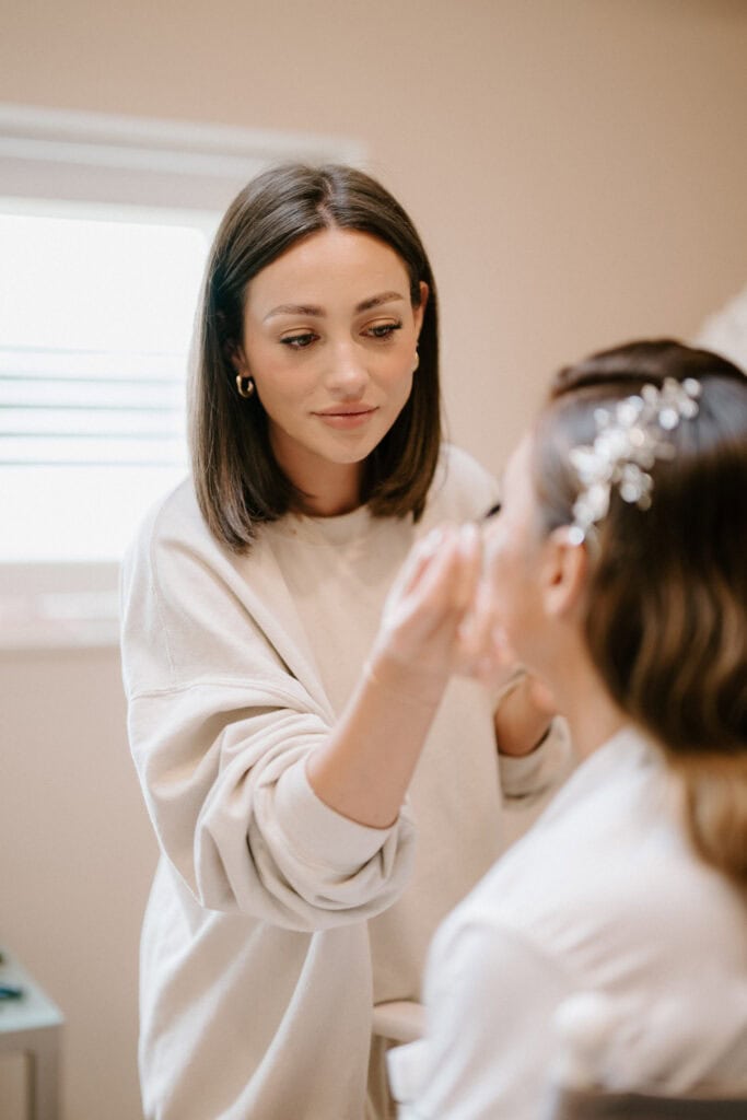 A woman with shoulder-length brown hair and a beige sweater is meticulously applying makeup to another individual's face, preparing them for a KENT WEDDING at Winters Barns. The person receiving makeup has styled hair adorned with decorative hairpins, in a well-lit room with a window in the background. Image by Pearce Wedding Photography.