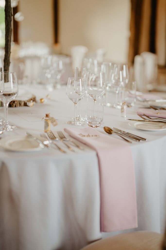 Elegant table setting with white tablecloths, pink napkins, and polished silverware at Winters Barns. Crystal wine glasses and goblets are neatly arranged, exuding a sense of grace befitting a Kent wedding celebration. Image by Pearce Wedding Photography.