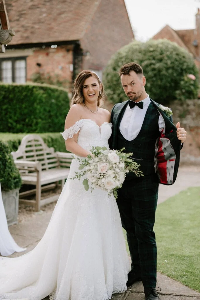A bride and groom stand outdoors at Winters Barns. The bride, wearing an off-the-shoulder lace wedding gown and holding a bouquet, smiles cheerfully. The groom, in a checkered suit with a bow tie, strikes a playful pose by showing the inside of his suit jacket. They are in a garden setting at this picturesque Kent wedding. Image by Pearce Wedding Photography.