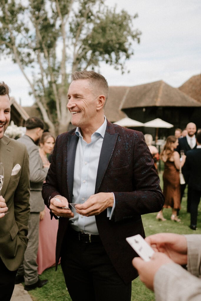 A man wearing a dark blazer and light blue shirt smiles while holding cards in an outdoor setting at a KENT WEDDING. He is surrounded by people, some holding drinks, at the Winters Barns social event with trees and buildings in the background. Image by Pearce Wedding Photography.