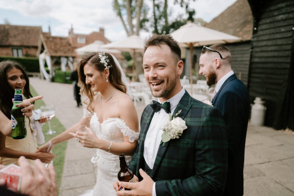 A smiling groom in a green plaid suit with a bow tie and floral boutonniere stands outdoors at Winters Barns, holding a beer bottle. A bride in a white, off-shoulder wedding dress holds a drink beside him. Other guests are mingling in the background near umbrellas and buildings at this charming Kent wedding. Image by Pearce Wedding Photography.