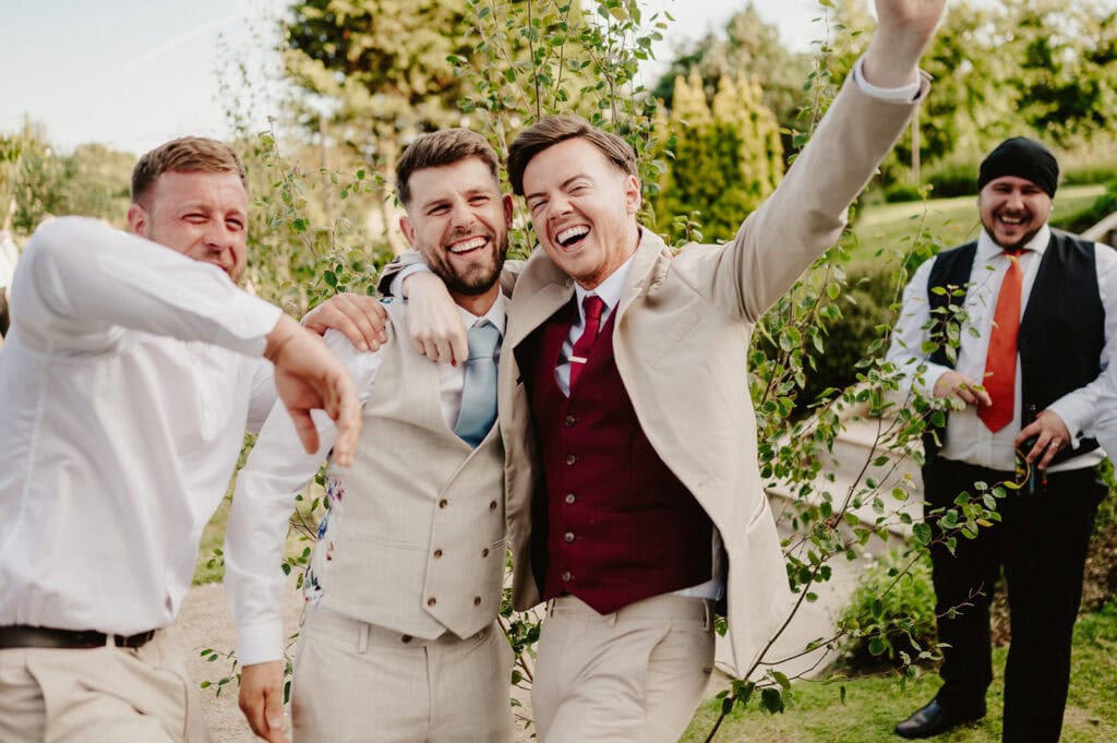 Four men at an outdoor event, dressed in formal attire. Three of them are laughing and posing closely for a photo, while the fourth, in the background at Reach Court Farm, is smiling and holding a drink. Trees and greenery are visible in the background, indicating an outdoor setting captured by a Dover wedding photographer. Image by Pearce Wedding Photography.