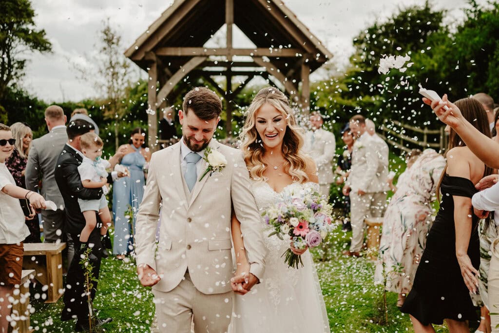 A bride and groom walk hand in hand, smiling, under a wooden arch at Reach Court Farm as confetti is thrown by guests. The bride holds a bouquet of flowers, and both are dressed in wedding attire. Guests on either side capture the moment on their phones and cameras, perfect for any Dover wedding photographer. Image by Pearce Wedding Photography.