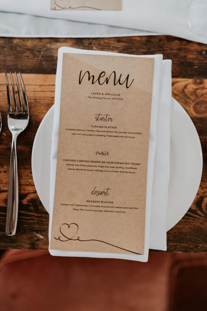 A wedding menu at The Winding House on a rustic wooden table setting features a fork on the left and a knife on the right. The menu offers a Turkish platter for starters, chicken carving board or vegetarian nut roast for the main course, and a dessert platter for dessert. Image by Pearce Wedding Photography.