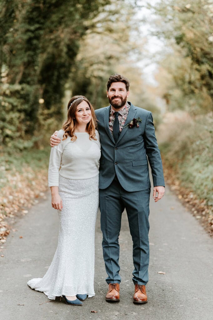 A couple stands on a tree-lined path. The woman, dressed in a light-colored, long skirt and top, stands beside a bearded man in a blue suit with a floral tie. Both are smiling, with the man's arm around the woman's shoulder. Captured by their Dover wedding photographer, the background is filled with greenery. Image by Pearce Wedding Photography.