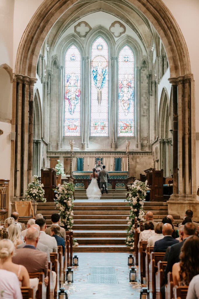 A bride and groom hold hands at the altar of St. Leonard's Church, a grand, ornate building in Hythe with high arches and large stained glass windows. The pews are filled with guests, and the aisle is adorned with flowers. Bathed in natural light, they stand on steps leading up to the altar as their wedding photographer captures every moment. Image by Pearce Wedding Photography.
