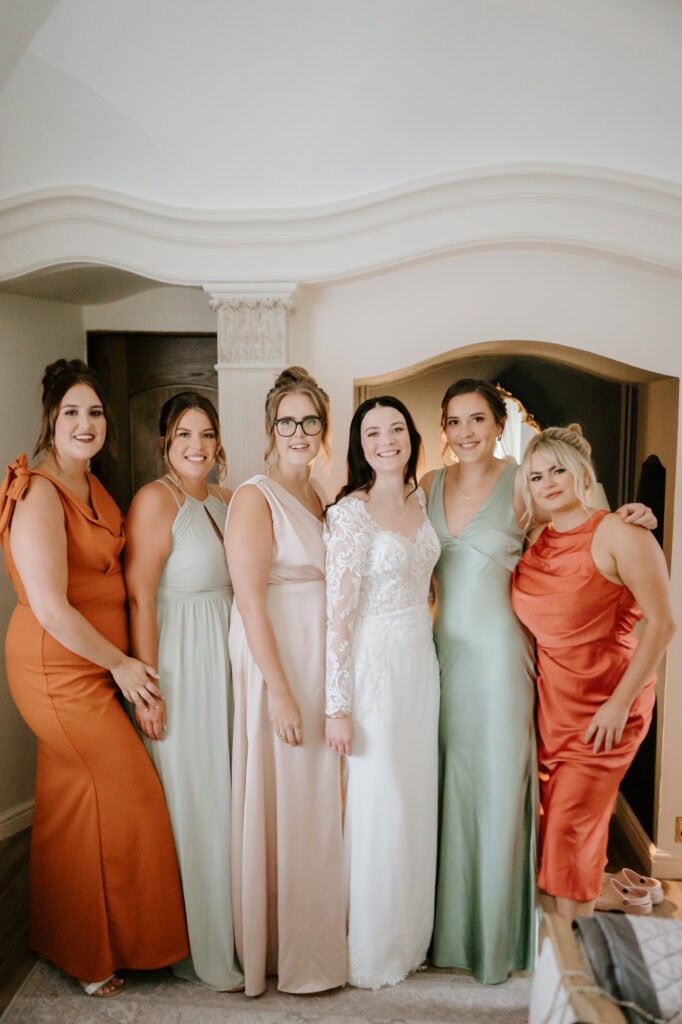 A bride, dressed in a long-sleeved lace wedding gown, stands smiling with five bridesmaids. The bridesmaids are wearing dresses in varying shades of green and orange. They are standing in a warmly lit room with an arched doorway, captured perfectly by a Hythe wedding photographer at Hayne House. Image by Pearce Wedding Photography.