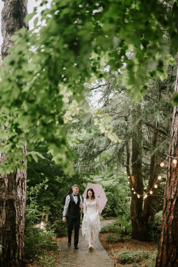 A couple walks along a forested path on a rainy day. The woman, wearing a white wedding dress, holds a white umbrella with red polka dots. The man, in a vest and tie, holds her hand. String lights hang from the trees, creating a romantic atmosphere captured beautifully by Hythe wedding photographer at Hayne House. Image by Pearce Wedding Photography.