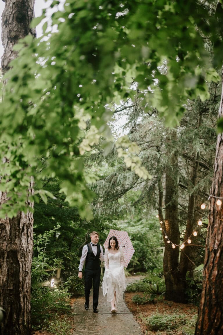 A couple walks along a forested path on a rainy day. The woman, wearing a white wedding dress, holds a white umbrella with red polka dots. The man, in a vest and tie, holds her hand. String lights hang from the trees, creating a romantic atmosphere captured beautifully by Hythe wedding photographer at Hayne House. Image by Pearce Wedding Photography.