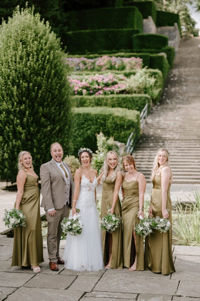 A bride in a white gown and groom in a beige suit stand outdoors with four bridesmaids in matching olive green dresses, all smiling and holding bouquets. Captured by a Hythe wedding photographer, they are framed by the manicured gardens and stone stairs at the scenic Port Lympne. Image by Pearce Wedding Photography.
